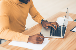 black man in orange shirt sitting and working on a written document and a laptop
