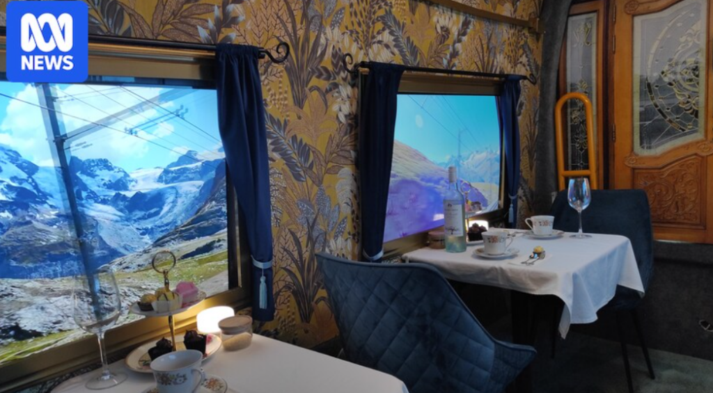 Dining setup inside a train carriage with a scenic mountain view displayed on the window, featuring elegant tableware, a glass of wine, and decorative elements, highlighting a unique dining experience related to travel and leisure.